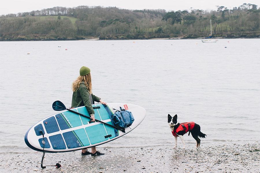 Woman carrying a loaded blue inflatable paddleboard into shallow water with a dog in a red buoyancy aid