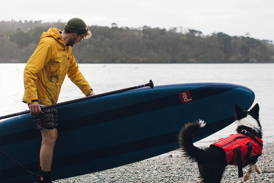 Man in a yellow jacket and beanie carrying a paddle board on a pebble beach with a dog in a red buoyancy aid