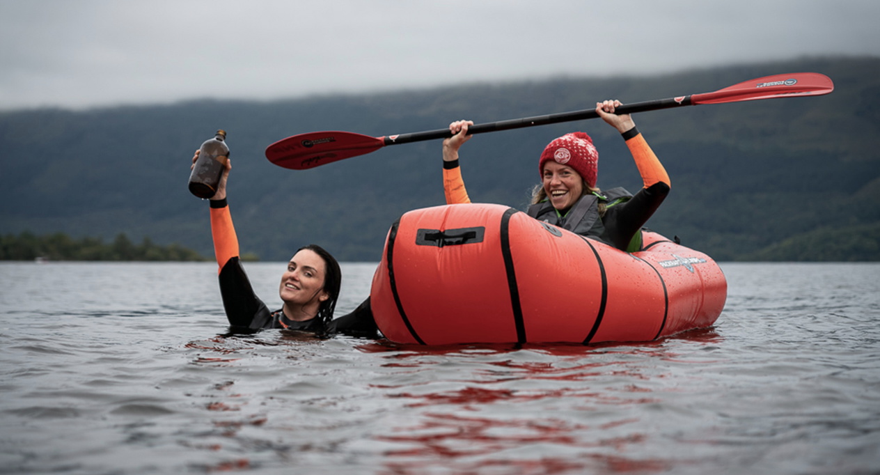 Two people in the cold open water beside a red inflatable packraft, one raising a paddle overhead