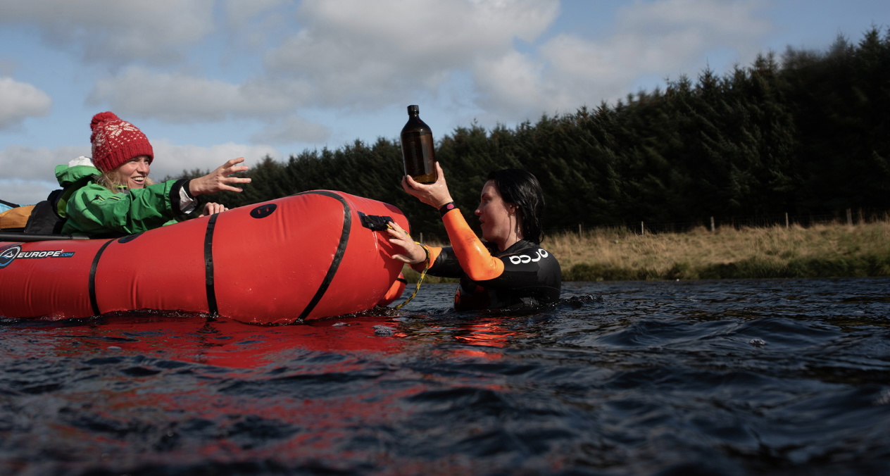 Two cold-water swimmers in a river sharing a hot drink beside a red inflatable packraft