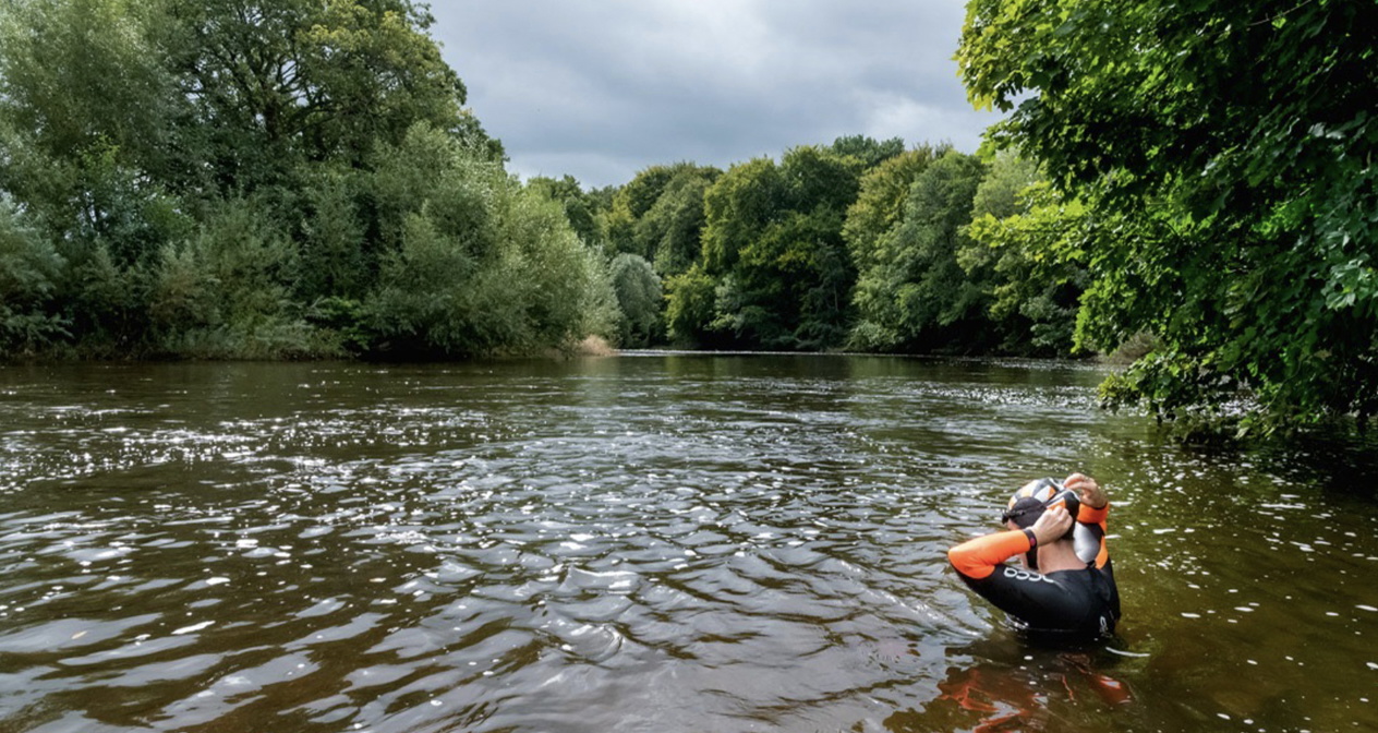 Wild swimmer wading into a green river with an orange tow float under a cloudy sky