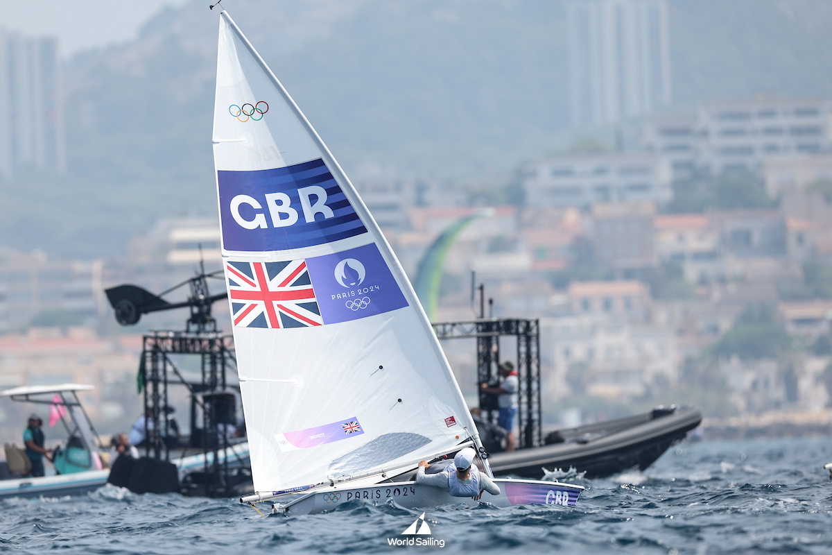Dinghy with a GBR sail carrying Olympic rings and a Union flag racing past a hazy coastal city