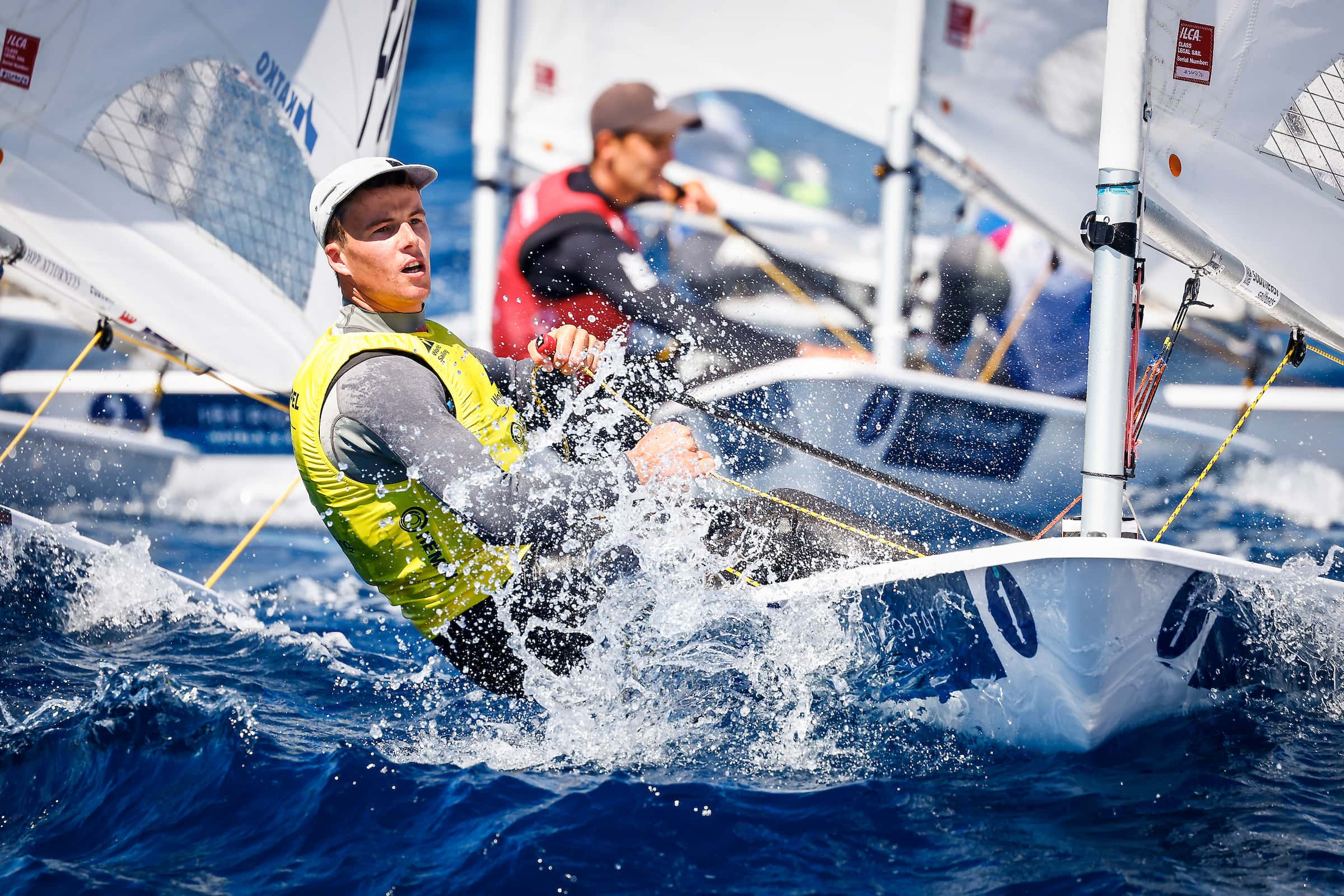 Sailor in a yellow bib hiking hard through heavy spray with rival boats close behind