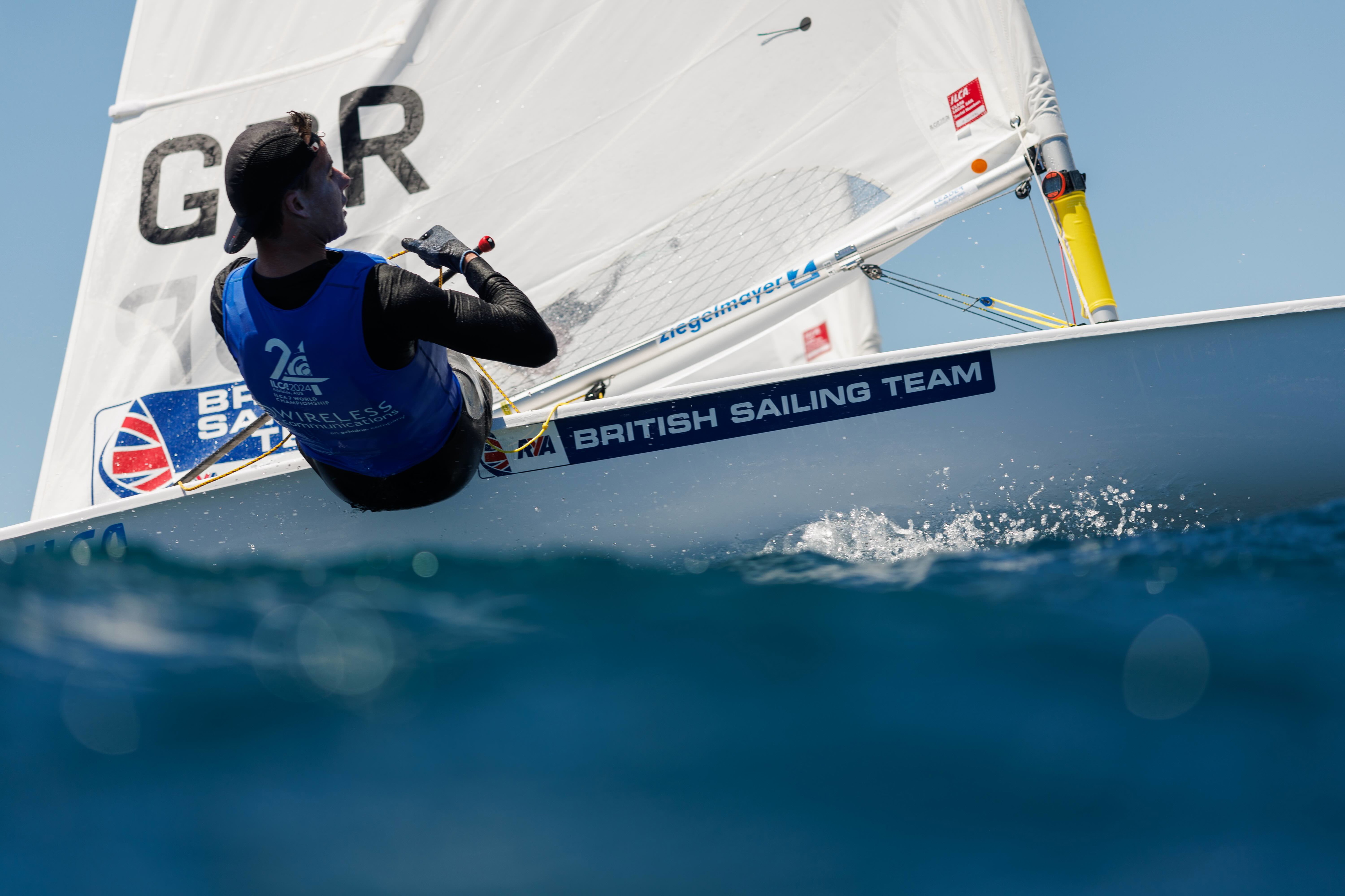 Sailor hiking out from a GBR-marked British Sailing Team dinghy on flat blue water
