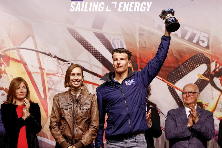 Sailor in a navy jacket raising a trophy on stage at a prizegiving as guests applaud
