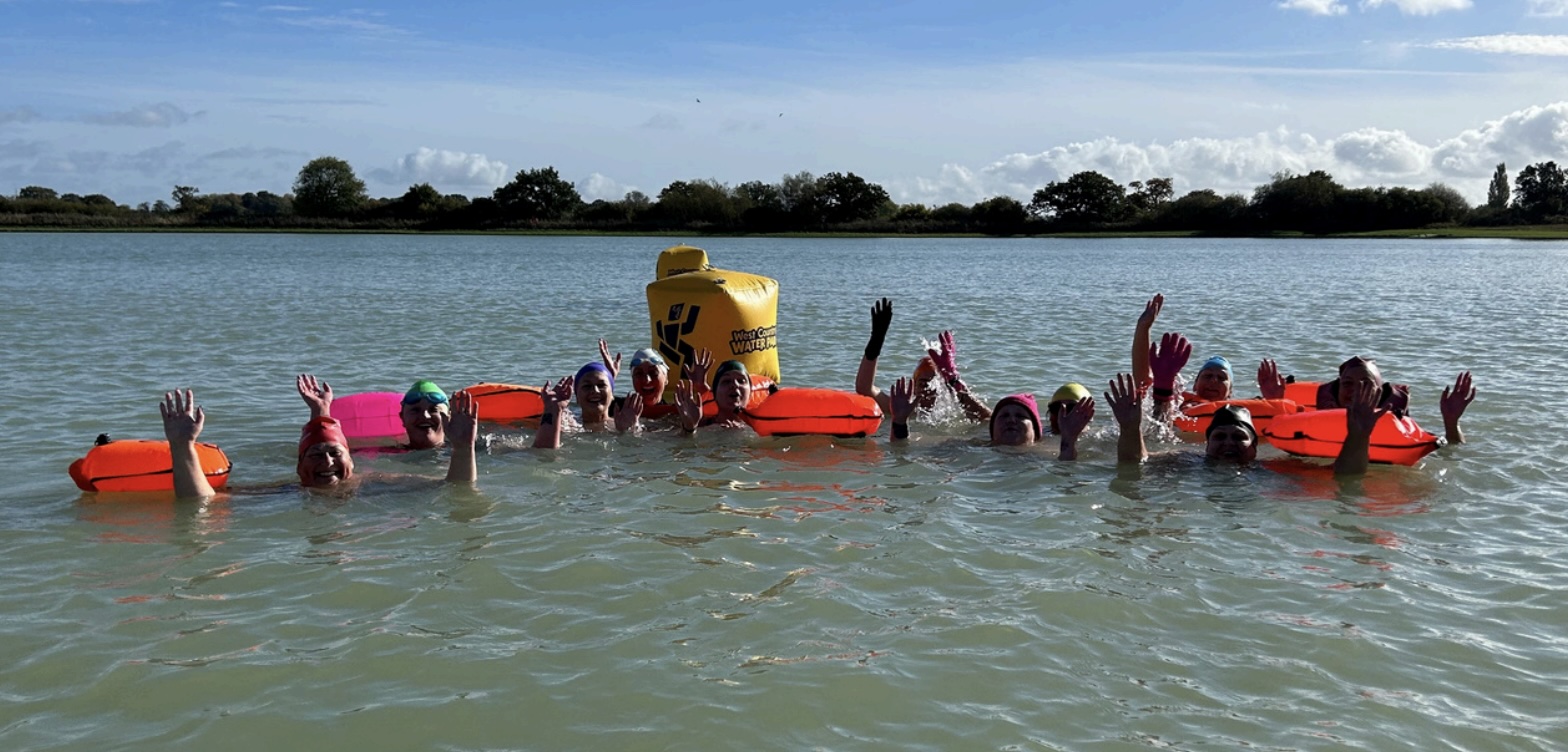 Large group of swimmers with orange tow floats raising their arms around a yellow buoy in a lake