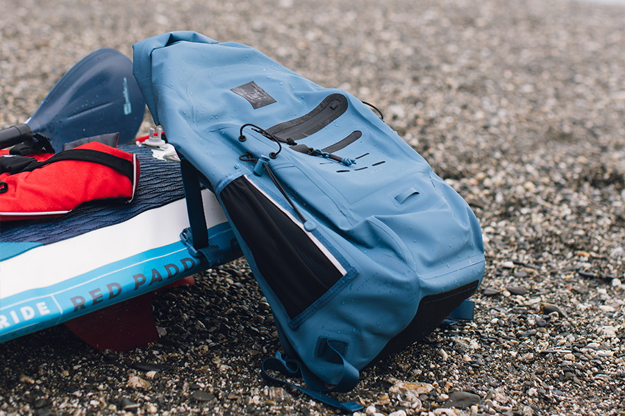 Blue roll-top dry backpack and a red buoyancy aid resting on a Red Paddle inflatable board on shingle
